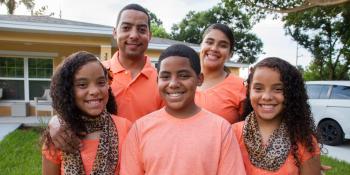 Family of five in matching orange shirts stand smiling in front of their home.