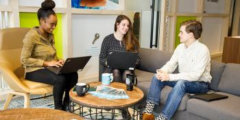 two women and a man chat together and work on laptops on couches and chairs in an office setting