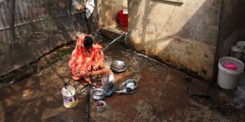 A woman in bright colored clothing washes dishes outside.