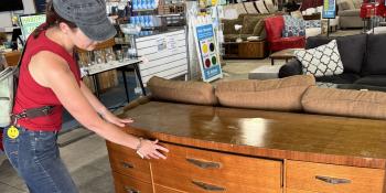 Woman in a red tank top, jeans and a gray hat inspecting a wood dresser.
