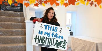 Photo of a smiling woman standing by a staircase, holding a hand-painted sign that says "This is my future Habitat home." There is an illustrated border of autumn leaves at the top of the image.