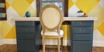Blue cabinet desk with a light wood desktop in front of a yellow wall with a light wood chair.