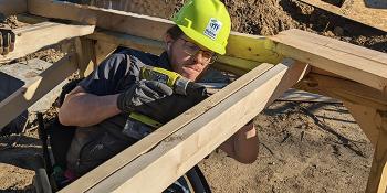 A young man wearing a yellow Habitat hardhat and using a wheelchair is drilling into wood boards on a home construction site.