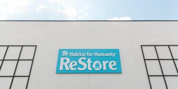 A photo of a white building and in the center is a blue sign that reads Habitat for Humanity ReStore and you can see blue skies and clouds above it.