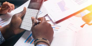 Two pairs of hands at a table. One is using a calculator. Printouts of charts are spread across the table.