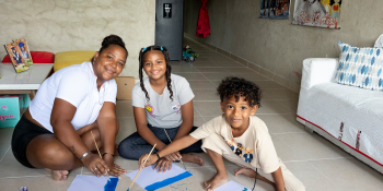 A mother sits on a ceramic tile floor with her two children while they paint sheets of paper blue. They're turned and smiling at the camera.