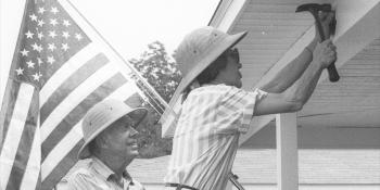 Black and white photo of President Carter watching Mrs. Carter hammer a beam on a Habitat home; an American flag is in the background, hanging from the house.