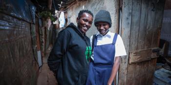 Two people standing in an alleyway, one is wearing overalls the other a blue sweatshirt.