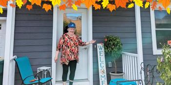 An older woman wearing a blue Habitat hardhat poses smiling in front of a display of photos and decor in her home. A graphic border of autumn leaves lines the top of the image.