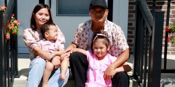 Sitting on the front stoop of their home, a mother and father hold their two young children, all in coordinating light pink outfits.