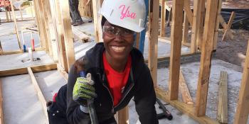 A woman wearing a hardhat with name 'Ysa' smiles as she is hammering wood framing on a Habitat build site