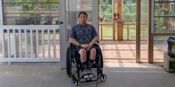 A young man in a wheelchair smiles next to a porch railing, with a greenhouse filled with plants behind him.