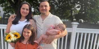 A family of four poses smiling together. The mother holds a bouquet of flowers, the father holds their baby, and their young daughter stands between them.