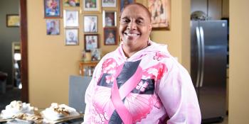 Woman in pink hoodie smiles in front of framed family photos in her home.