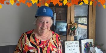 An older woman wearing a blue Habitat hardhat poses smiling in front of a display of photos and decor in her home. A graphic border of autumn leaves lines the top of the image.