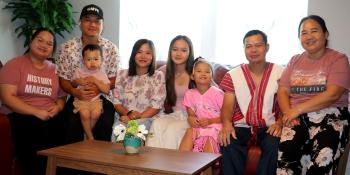 A multi-generational family with two women, two men, three young girls and a baby all pose together on a couch wearing matching pink-themed clothing