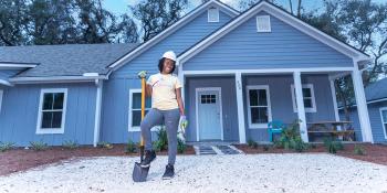 A woman wearing a hardhat smiles as she holds a shovel and poses in front of a new house.