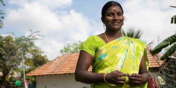 An Indian woman in green stands outside a tan-roofed house.