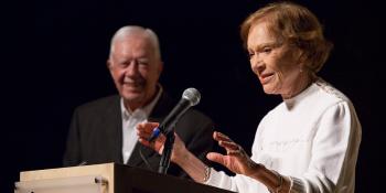 Mrs. Carter standing at a microphone on a podium with her hands raised as she talks to the audience. President Carter is standing to the side smiling at her.