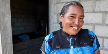 A woman in a blue shirt stands in front of her home, smiling.