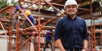 Man standing in front of construction site smiling at camera while wearing a hard hat