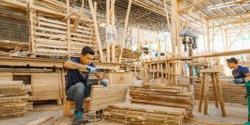 A man is sitting in warehouse surrounded by lumber and hammering a wooden pallet together.