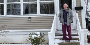 A man with gray hair standing on the porch steps in front of his beige home.