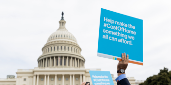 Picture of a sign being held up by a hand that reads, "Help make the #CostOfHome something we all can afford." with the Capitol Hill building in the background.