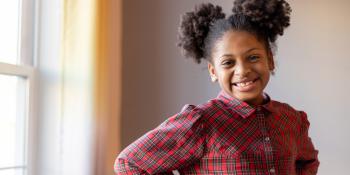 A young girl with pigtails stands by a window smiling.