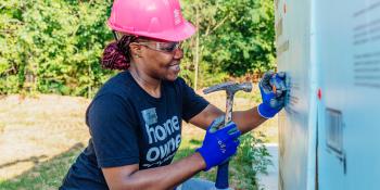 A woman wearing a pink helmet and a T-shirt that reads "home owner" smiles as she hammers.
