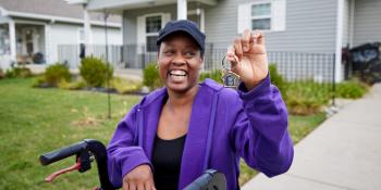 Aretha holds up a key, smiling as she sits on her walker in the driveway of her home.