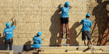 Four Habitat volunteers in bright blue shirts and hard hats hammer nails into a wall of plywood.