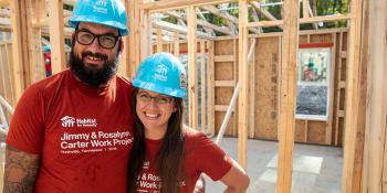 Neil and Erica stand together in blue hard hats, smiling at the camera in their soon-to-be home, which is lumber and beams at the time of the photo.