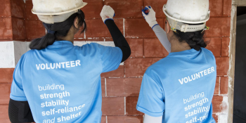 Two women paint a brick wall together. They are wearing white hardhats and the backs of their blue shirts say "Volunteer - building strength, stability, self reliance and shelter"