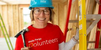 Woman in red Wells Fargo volunteer t-shirt on a construction site with a blue Habitat hardhat on holding a hammer.