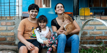 Woman sitting with her two children and holding a small dog. The family is smiling and they are framed by the vibrant blue wall of the house.