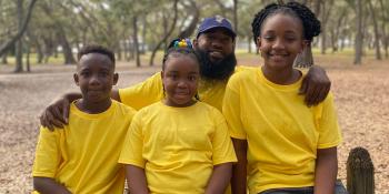 A father has his arms around his three children, an adolescent girl and a girl and boy of elementary school age. All of them are smiling in matching yellow shirts with trees in the background.