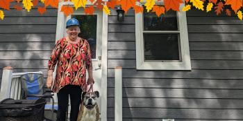An older woman wearing a blue Habitat hardhat poses smiling with her dog in front of her home. A graphic border of autumn leaves lines the top of the image.