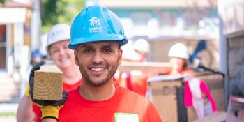 Man volunteer in blue Habitat hardhat and red Wells Fargo shirt carrying wood on Habitat build site