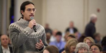Man wearing sweater is holding a mic and speaking in a crowded room.