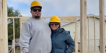 Couple wearing hardhats smile and pose in the framing of their future Habitat house.