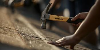 Close-up of hands hammering nails on a roof, with others hammering in the background.