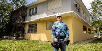 José with hard hat and tool belt in front of home repair site.