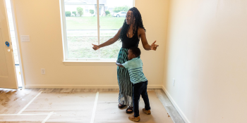 A woman is standing near a window holding her arms out as she looks around at the freshly painted living room of her new Habitat home. Her young son hugs her tightly around the waist.