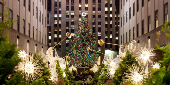 A very large decorated Christmas tree stands in Rockefeller Center plaza surrounded by smaller trees, star-shaped lights, and golden trumpets.