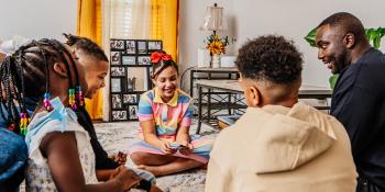 A father and his 4 kids sit on their living room rug playing a card game and laughing together