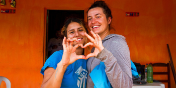 Two young women volunteers with Habitat shirts smile and pose with their hands making a heart shape.