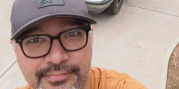 Man in an orange shirt, glasses and gray hat in front of a silver truck.