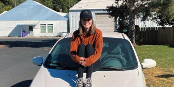 woman in an orange jacket and a black cap sitting on the hod of a white car.