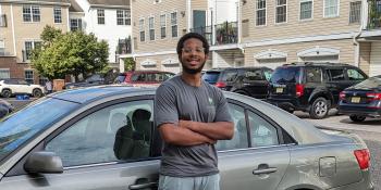 Gray car with a man in shorts, flip flops and a gray t-shirt standing next to it.